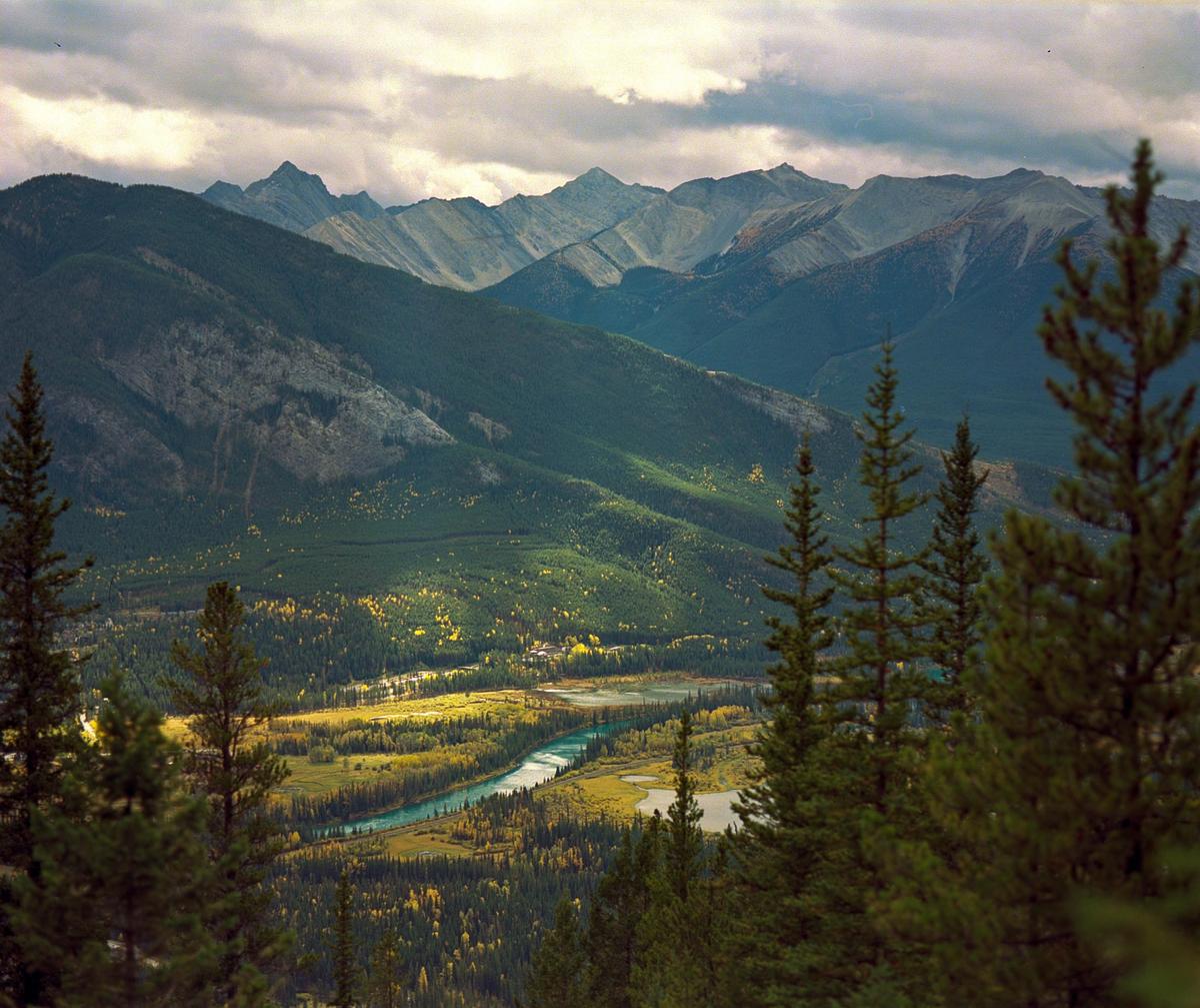 A river winds through a valley with autumn foliage, surrounded by mountains under a cloudy sky.