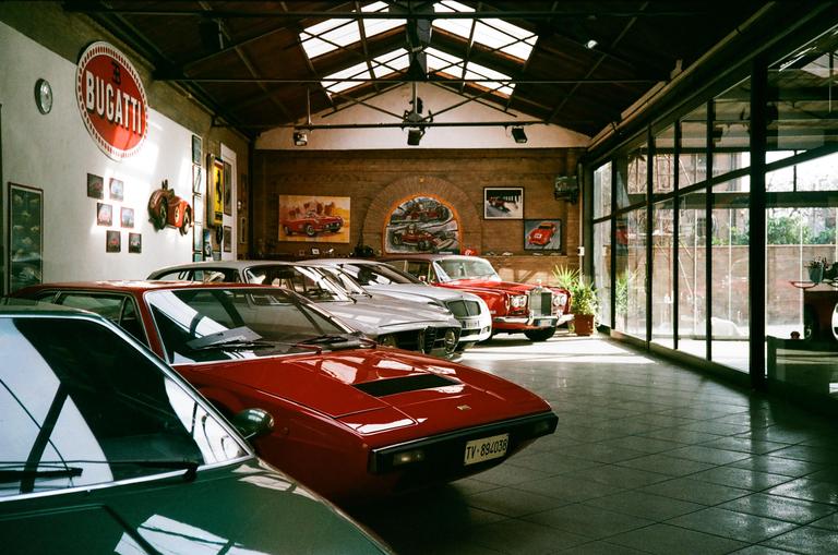 A showroom with several vintage cars, including a red sports car in the foreground, is illuminated by natural light from skylights and large windows.