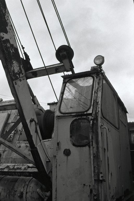 A black and white photograph shows the weathered cabin and boom of a large piece of industrial machinery against a cloudy sky.