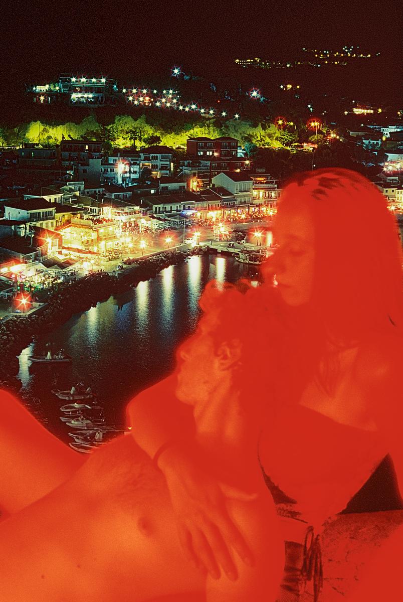 A couple is embracing in the foreground with a brightly lit town and harbor visible in the background at night.