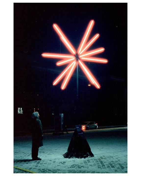A person stands on a snowy street at night, looking up at a large neon star sculpture.