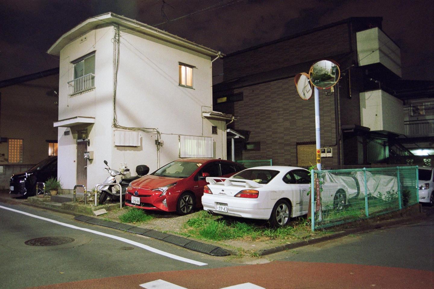 A white house with illuminated windows stands next to a street with parked cars and a scooter at night.
