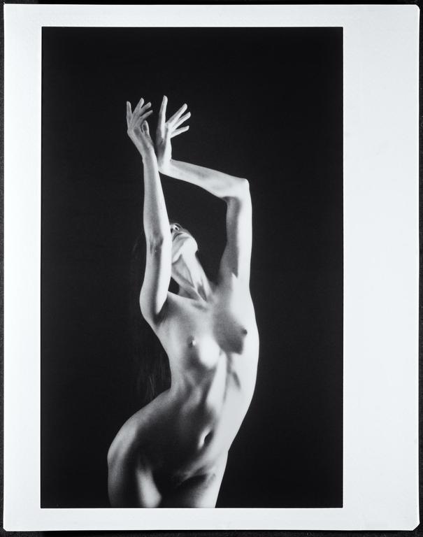 A nude woman with her arms raised and head tilted back is captured in a studio setting.