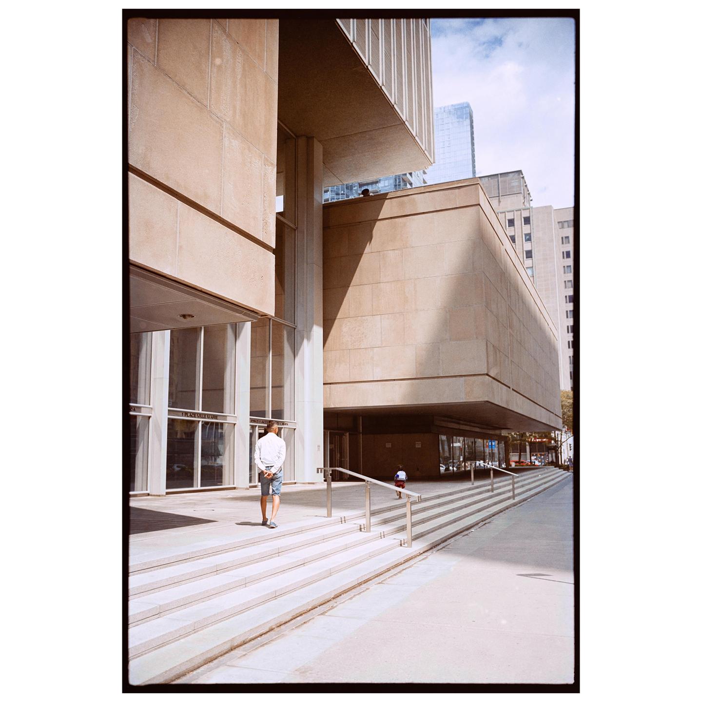 A man walks up a wide set of stairs towards a large concrete building with glass entrances.