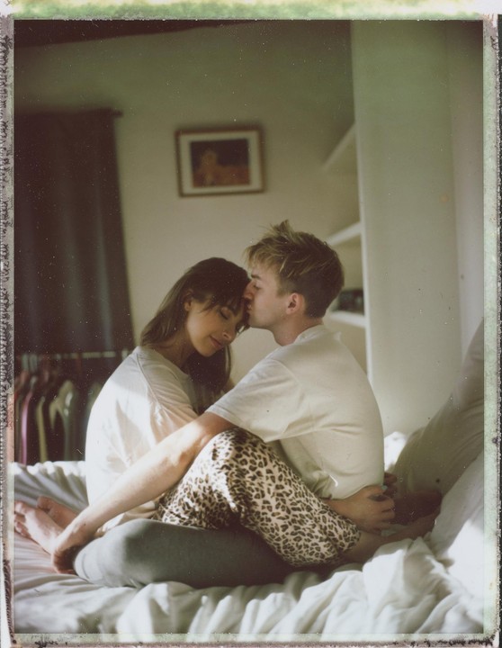 A man kisses a woman's forehead as they sit on a bed.