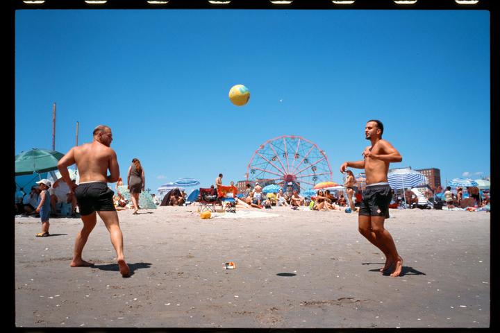 Two men play with a beach ball on a crowded beach with a ferris wheel in the background.