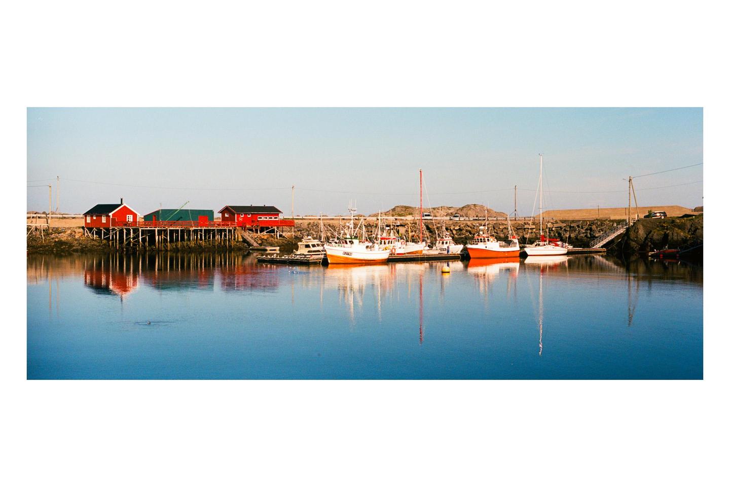 Several boats are docked in a calm harbor with red buildings on stilts along the shore.