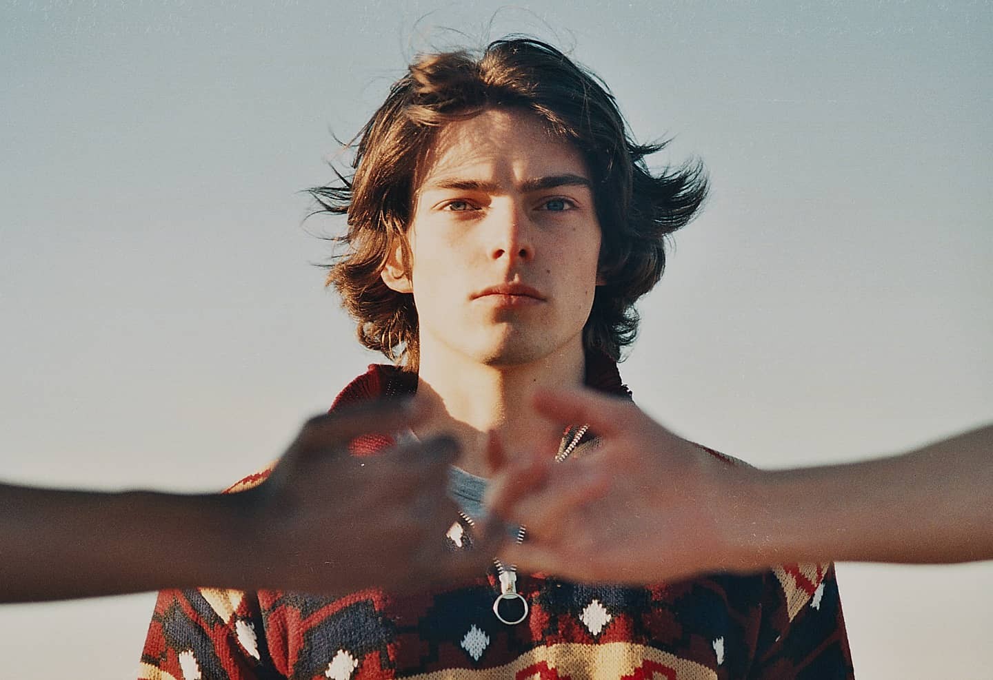 A man with long hair and a patterned sweater looks directly at the viewer, with two hands in the foreground forming a heart shape.