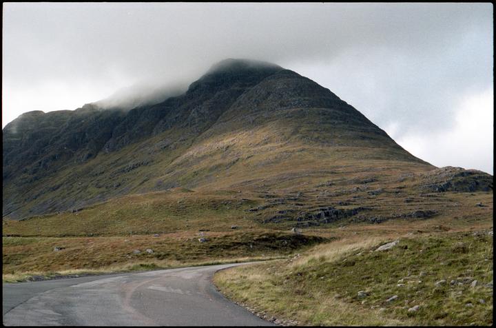 A winding road leads towards a large mountain partially obscured by clouds.
