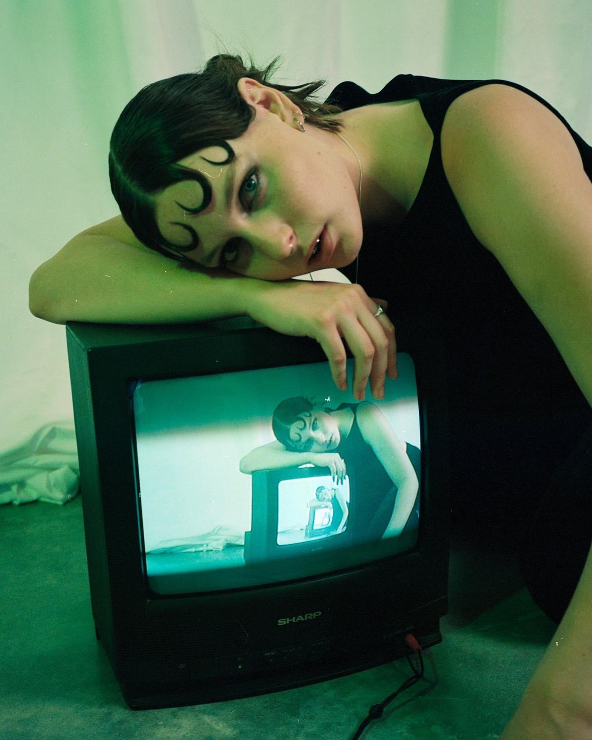 A woman with dark hair and a black top leans on a television set, which displays a recursive image of herself.