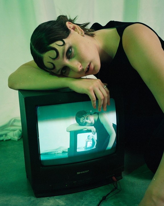 A woman with dark hair and a black top leans on a television set, which displays a recursive image of herself.