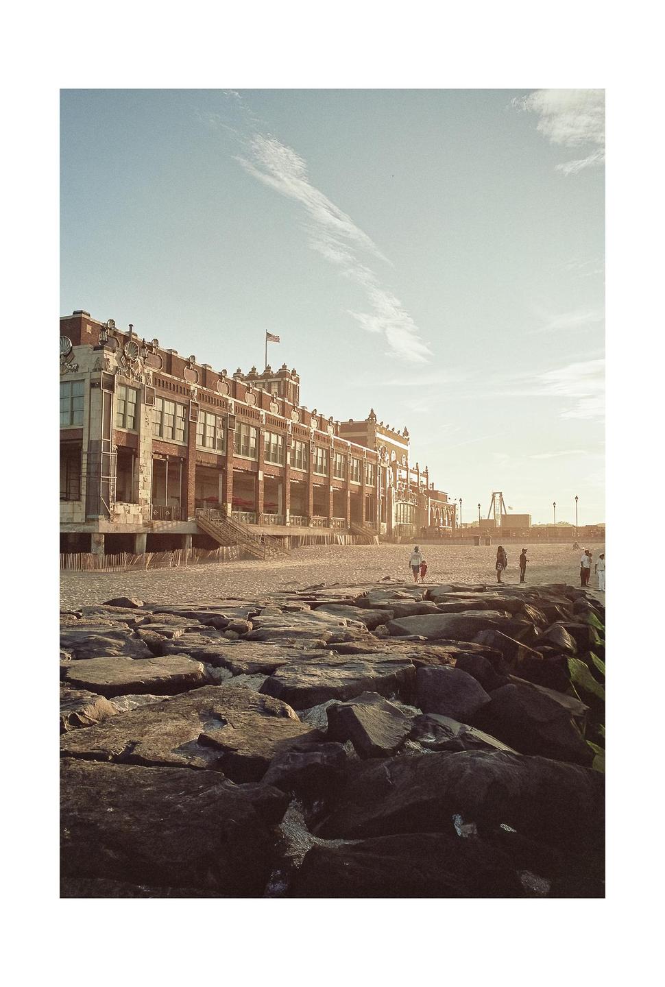 A large brick building with an American flag on its roof stands next to a sandy beach with rocks in the foreground and people in the distance.