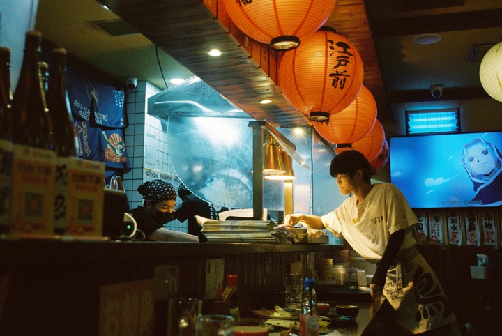 Two men are working behind a counter in an izakaya with orange lanterns hanging from the ceiling.