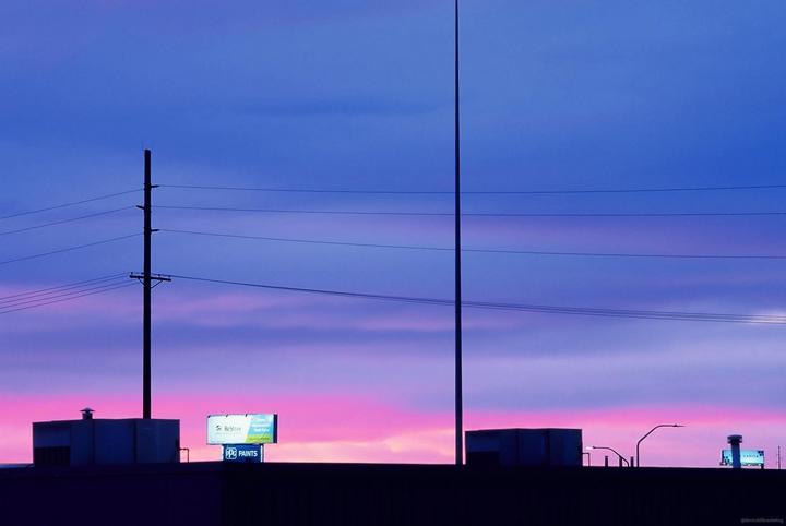 A vibrant pink and blue sunset sky is visible above a dark silhouette of buildings, power lines, and billboards.