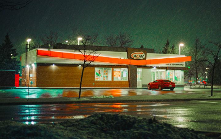 A red car is parked in front of an A&W restaurant at night while it is snowing.