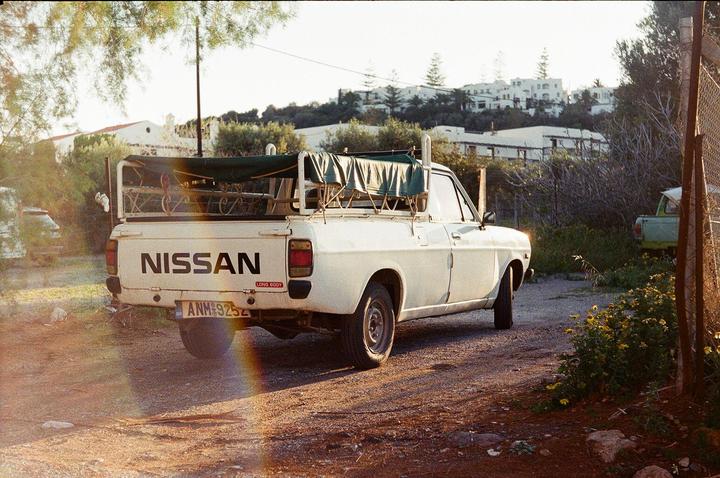A white Nissan pickup truck is parked on a dirt road with a village on a hill in the background.