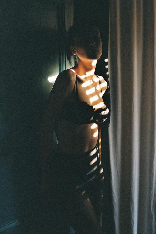 A woman stands in a dark room with stripes of sunlight falling on her body.
