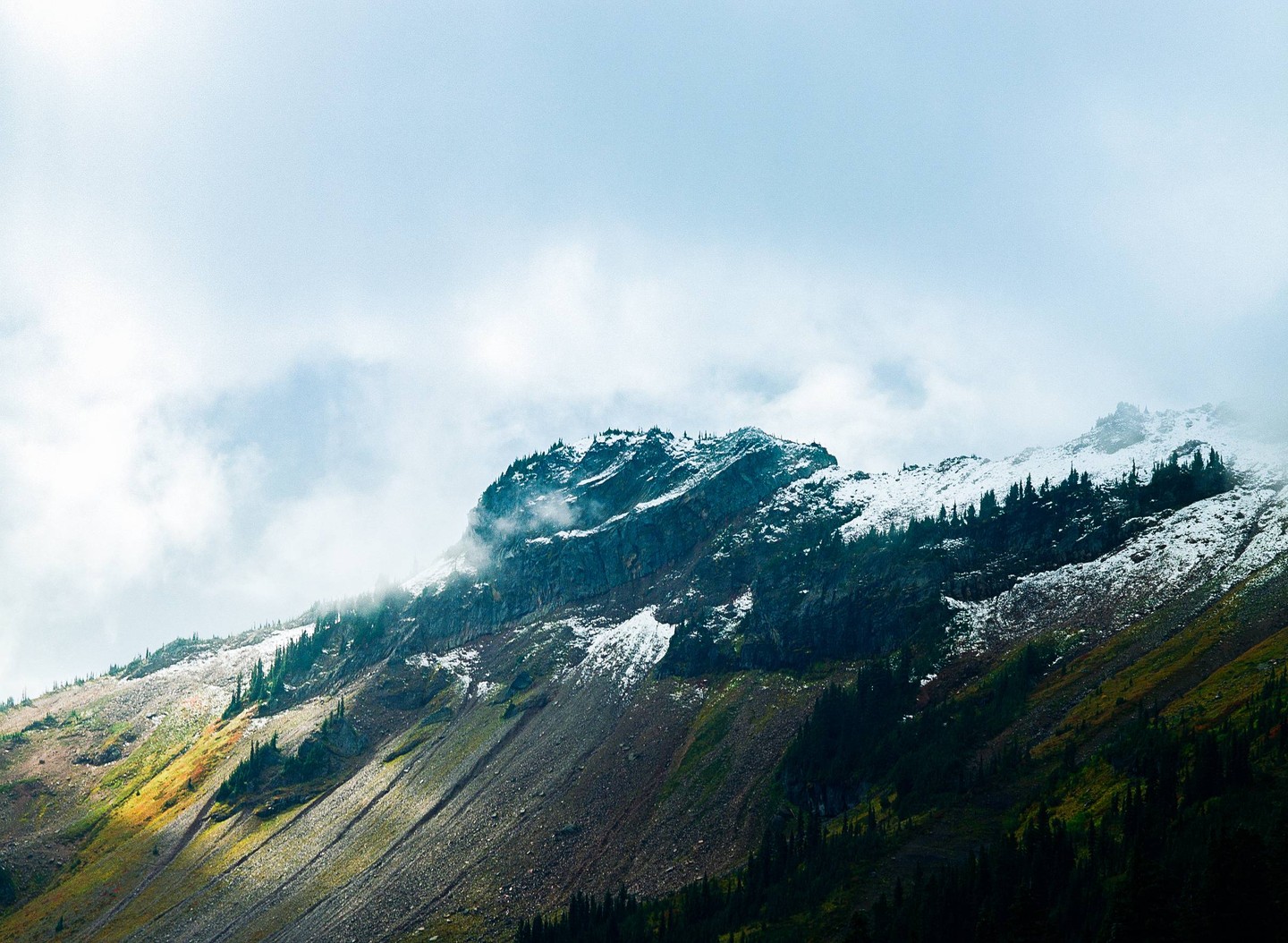 Clouds hang over a snow-capped mountain with patches of green and brown foliage.