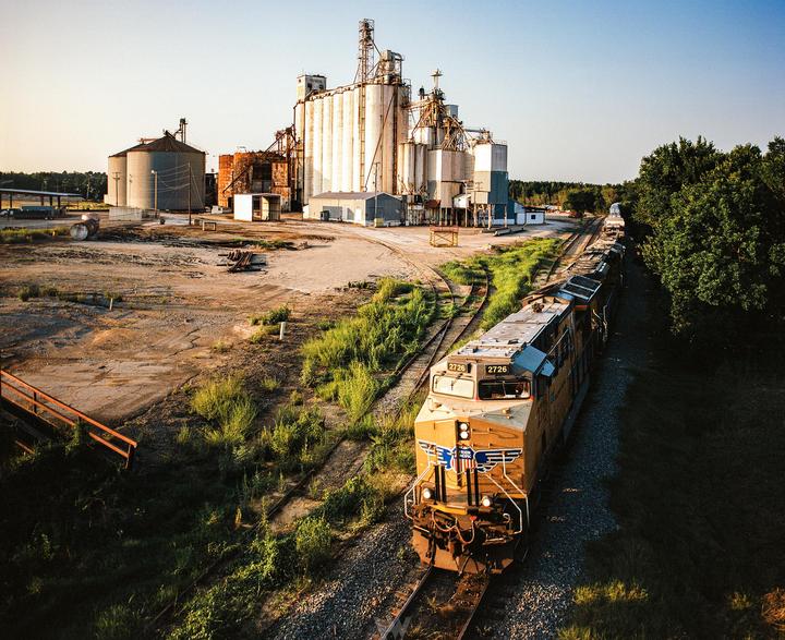A long train is parked on tracks next to a large industrial complex with silos and other buildings.