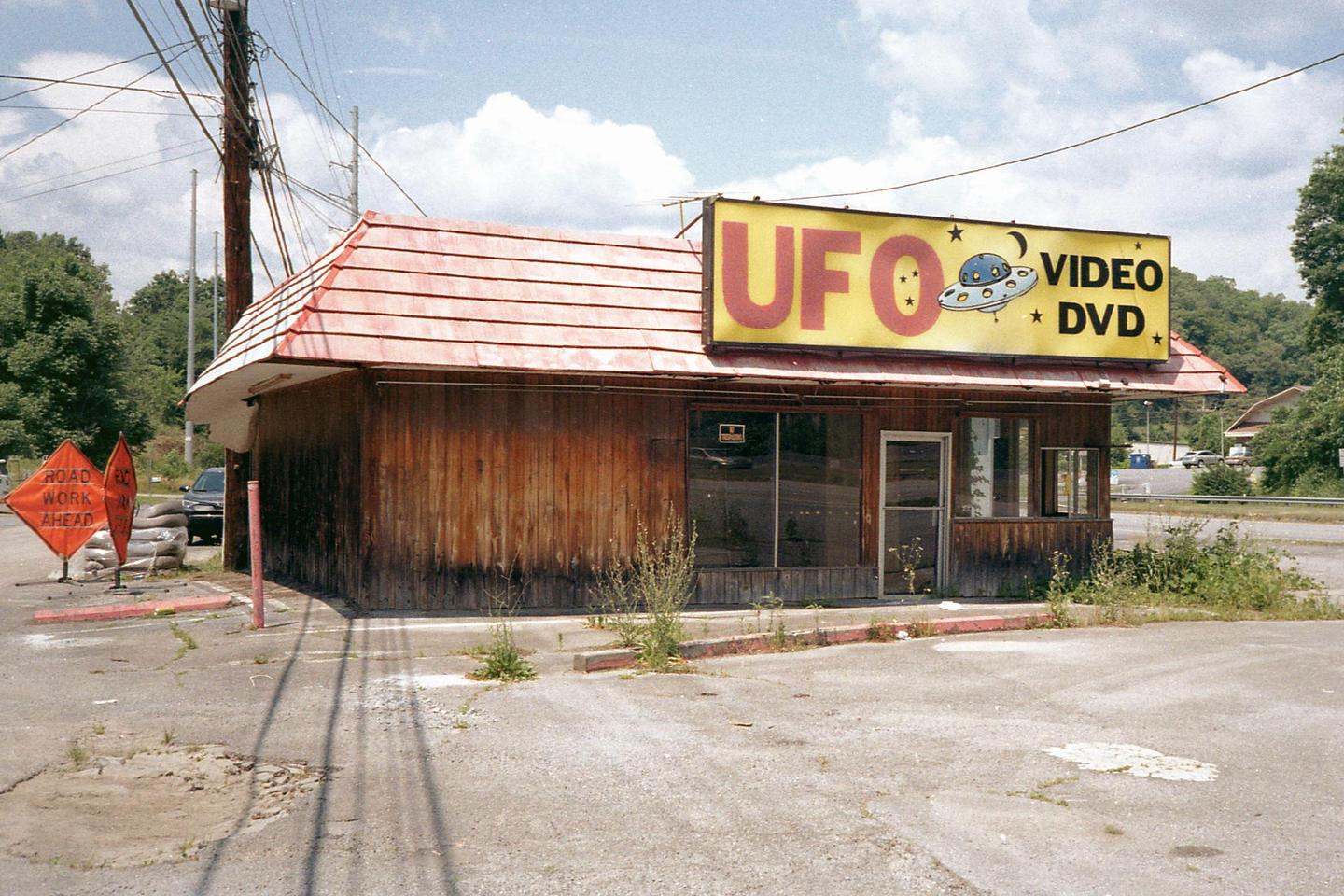 An abandoned UFO Video and DVD store with a red roof and wooden siding stands next to a road with "road work ahead" signs.