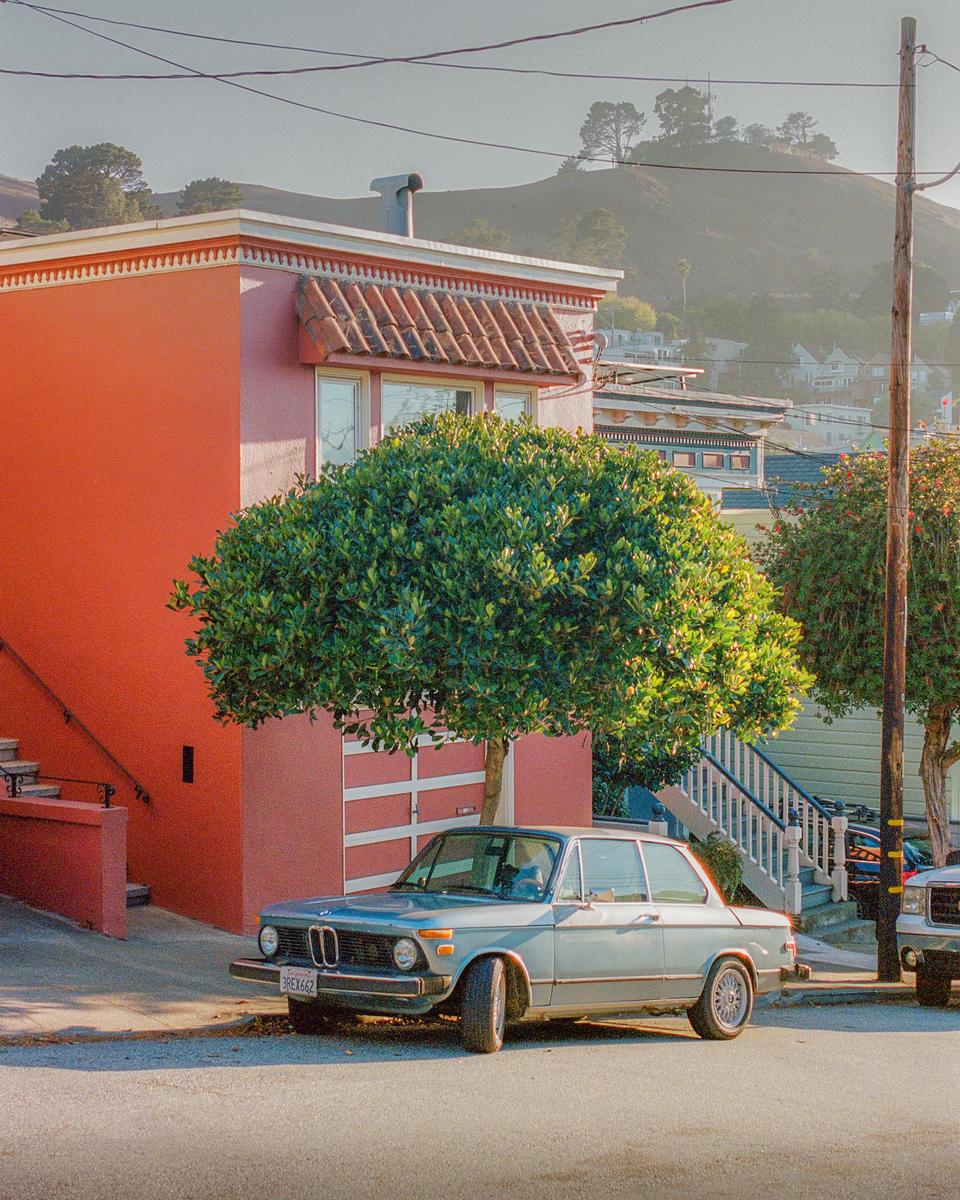 A light blue car is parked on a street in front of a red building with a green tree.
