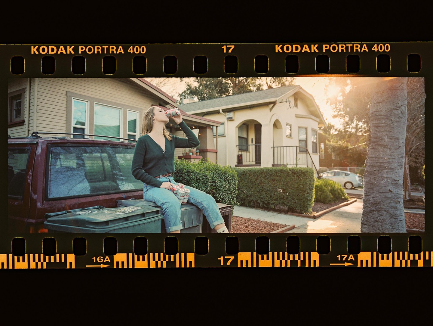 A woman sits on the back of a car, drinking from a can, with houses and a tree in the background.