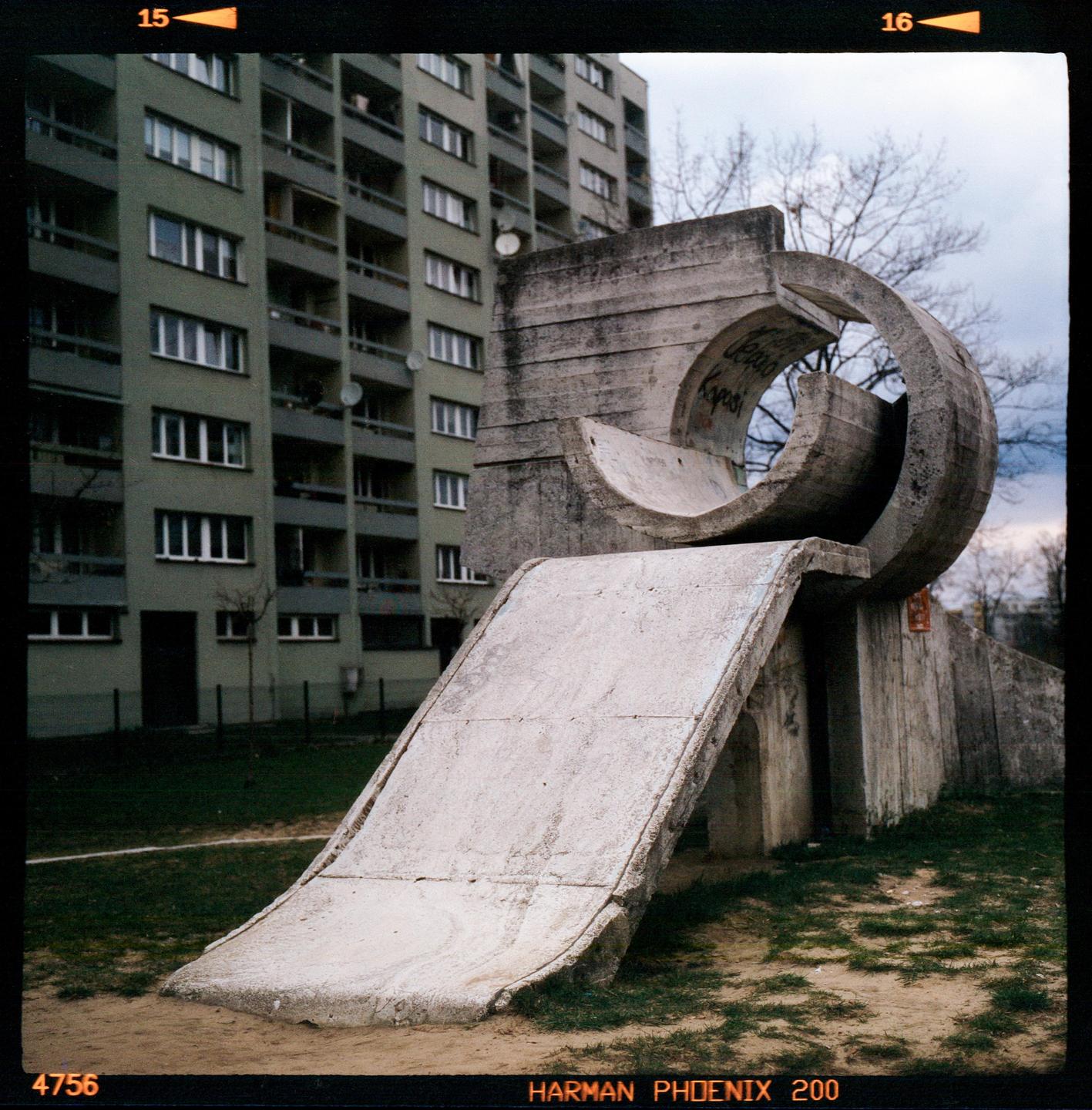 A concrete slide and abstract concrete structure stand in a grassy area in front of a large apartment building.