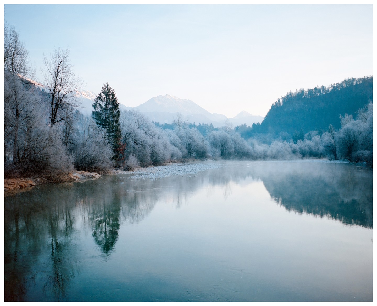 A wide river reflects a winter landscape with frost-covered trees and distant mountains under a clear sky.