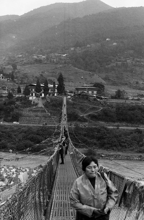 A woman walks across a long suspension bridge over a river, with mountains and buildings in the background.