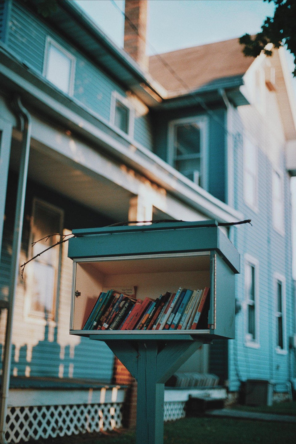 A small, light blue, outdoor library box filled with books stands in front of a house.