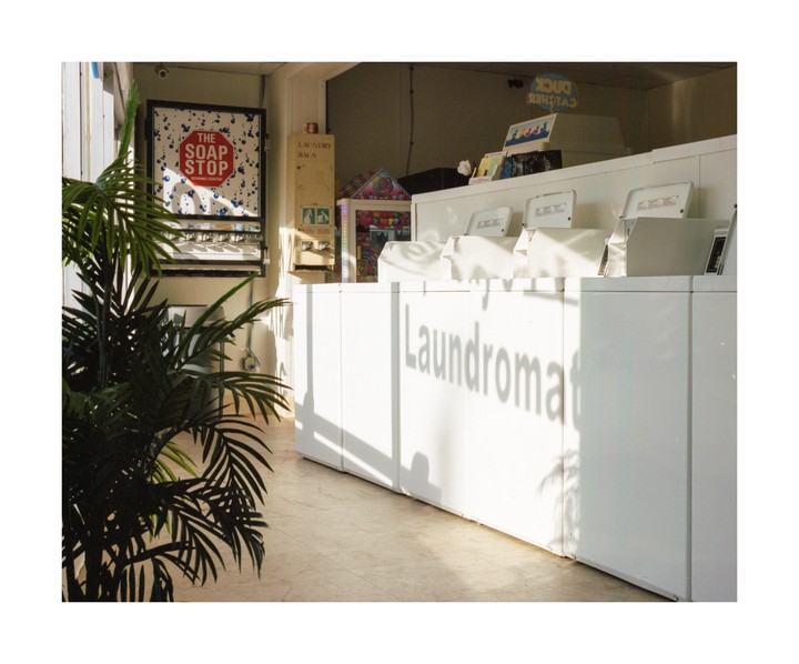 A laundromat interior features a "The Soap Stop" vending machine, a plant, and washing machines with "Laundromat" text and shadows cast upon them.