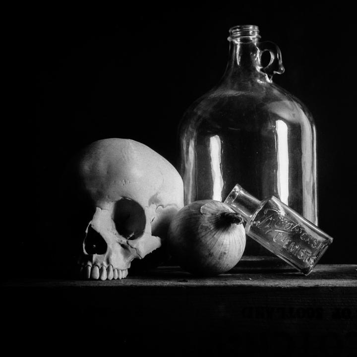 A still life arrangement features a skull, an onion, a large glass jug, and a small glass bottle on a wooden surface.