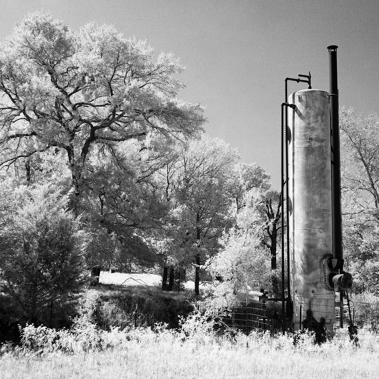 A tall industrial tank stands next to a field with trees in the background.