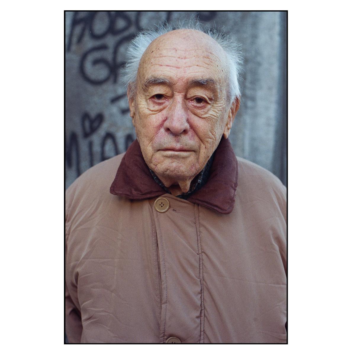 A man with white hair and a tan jacket stands in front of a wall with graffiti.