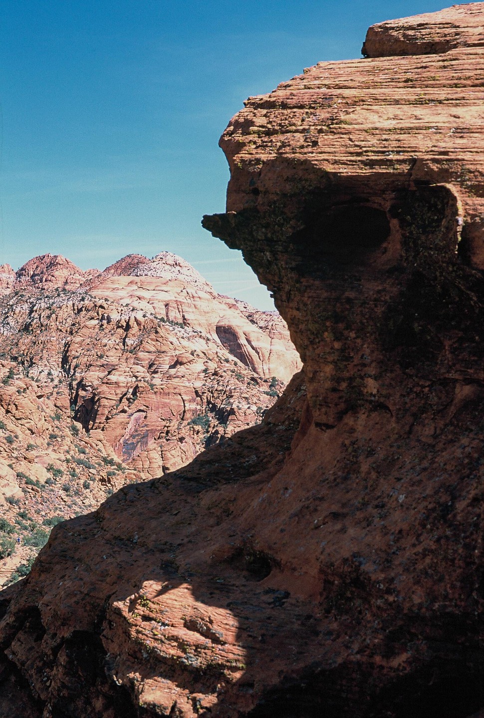 A large red rock formation with a profile resembling a face is visible in the foreground, with more red mountains in the background under a clear blue sky.