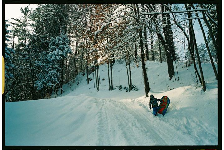 A child in winter clothing pulls a colorful sled up a snowy hill surrounded by trees.