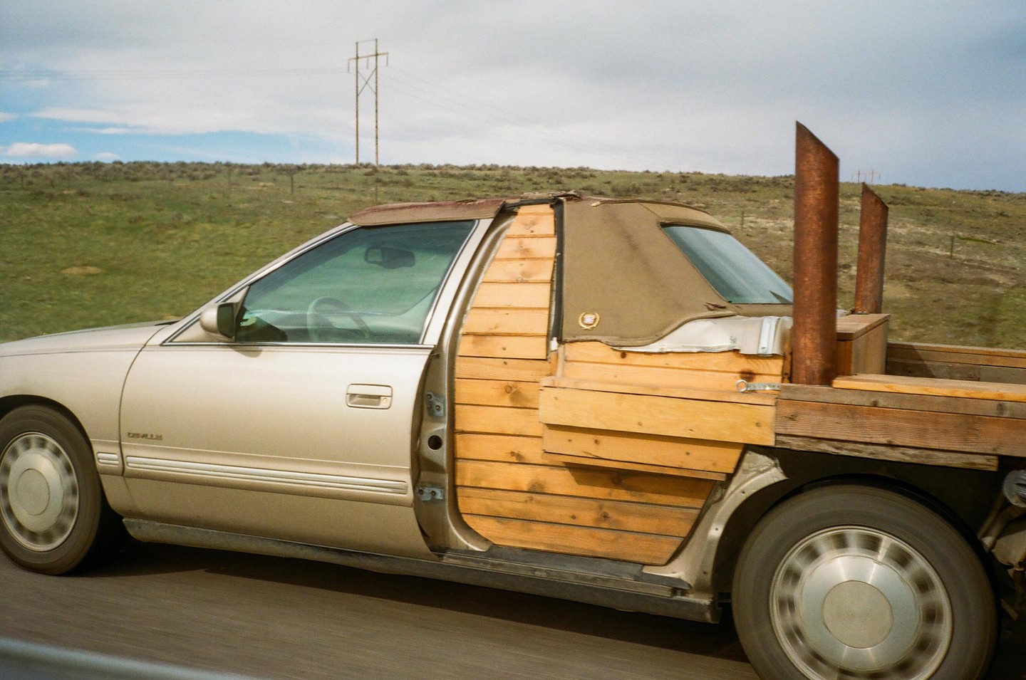 A gold car with a wooden flatbed and two tall exhaust pipes drives past a grassy hill under a cloudy sky.