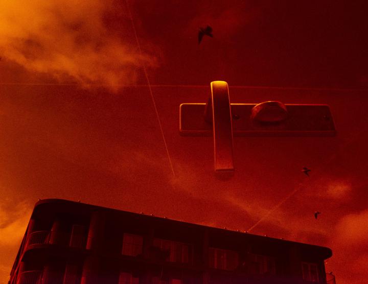 A building is visible below a door handle and birds flying in a red sky.