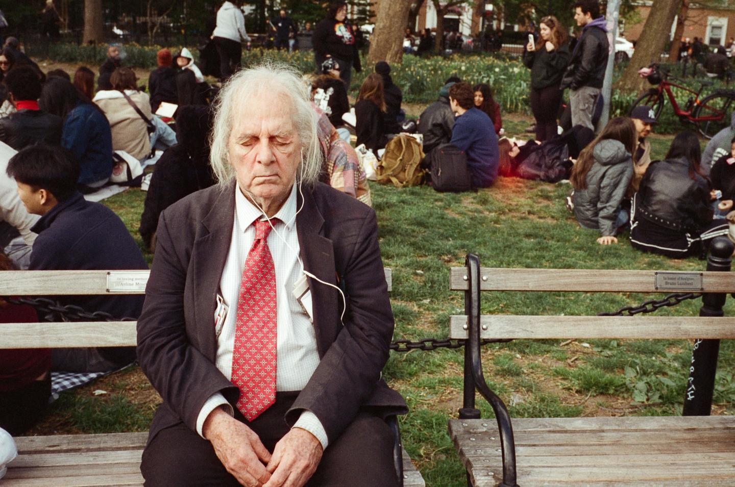 An elderly man with white hair and a red tie sits on a park bench with his eyes closed, wearing earphones, while other people relax on the grass in the background.