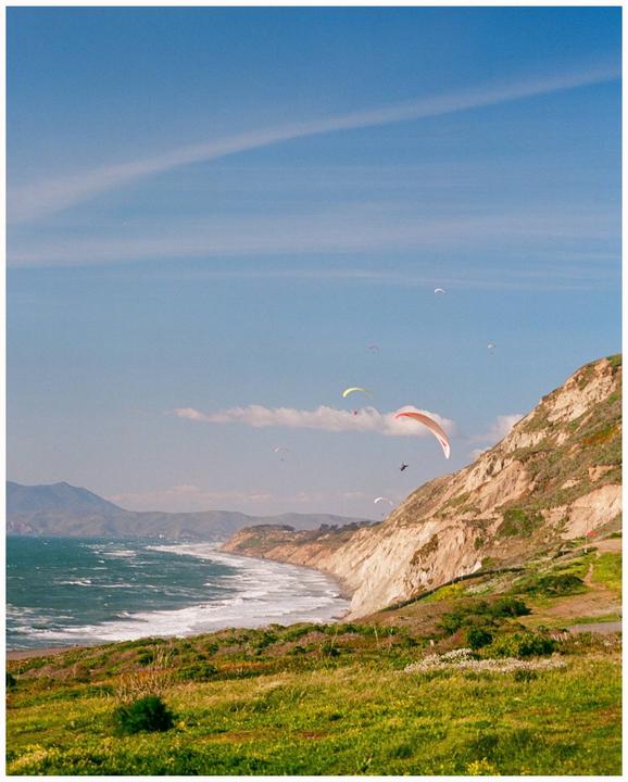 Several paragliders soar above a grassy cliff overlooking a wavy ocean and distant mountains.