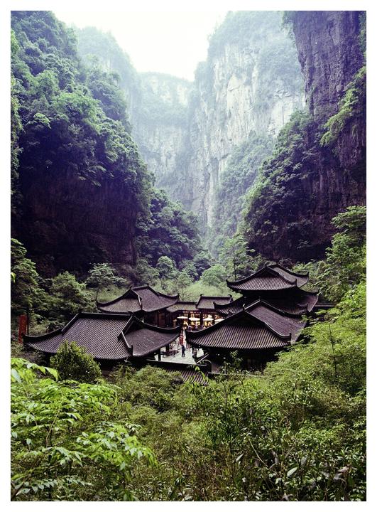 A cluster of traditional buildings with dark tiled roofs is nestled in a lush green valley surrounded by towering cliffs.