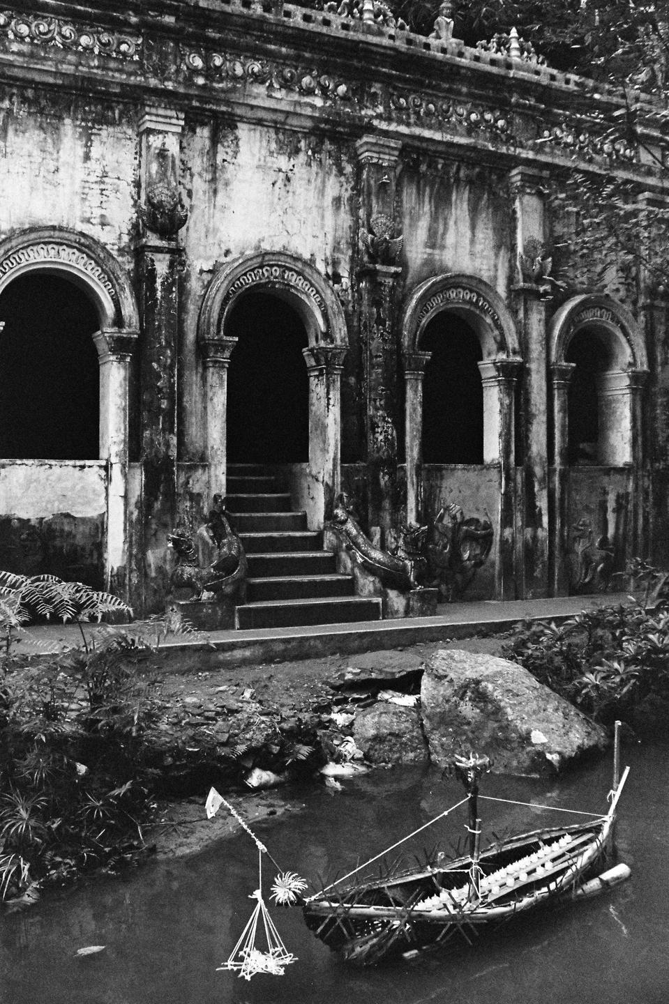 A small boat floats in a pond in front of an ornate, weathered building with arched doorways and a staircase.