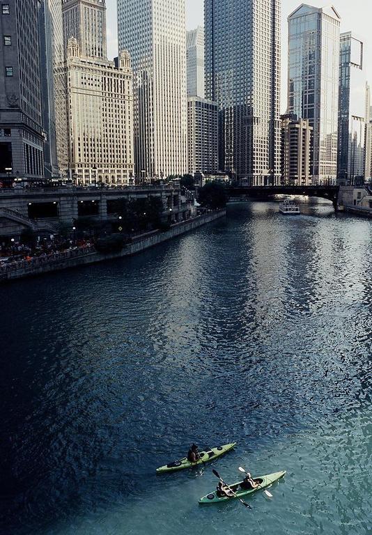 Two people kayak on a river in a city with tall buildings and bridges.