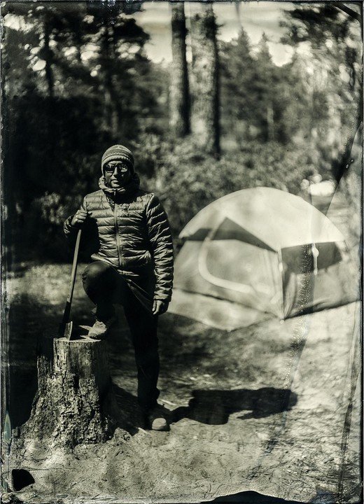 A man with an axe stands with one foot on a tree stump next to a tent in a forest.