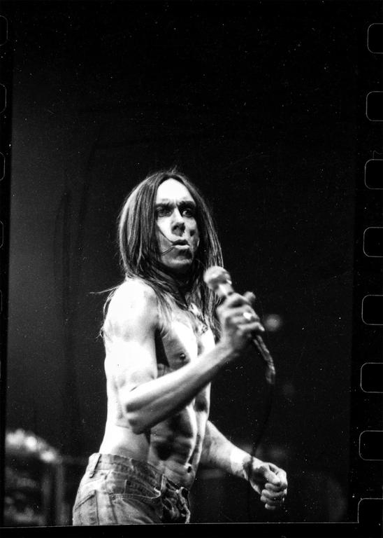 A shirtless man with long hair holds a microphone on a dark stage.