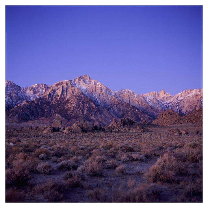 A vast desert landscape with sparse vegetation and large rocks leads to a snow-capped mountain range under a clear blue sky.
