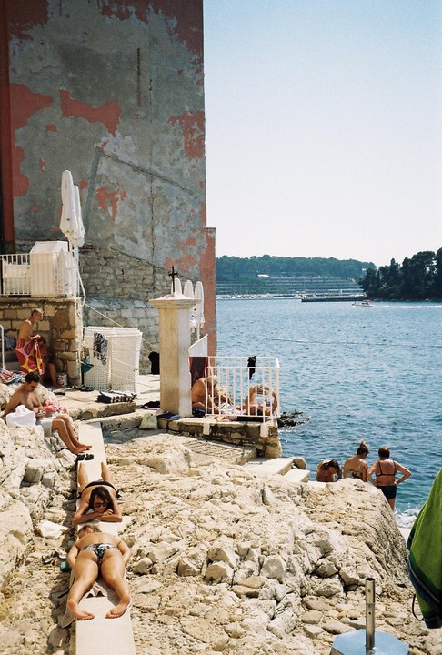 People sunbathe and swim on a rocky beach next to a building with peeling paint.