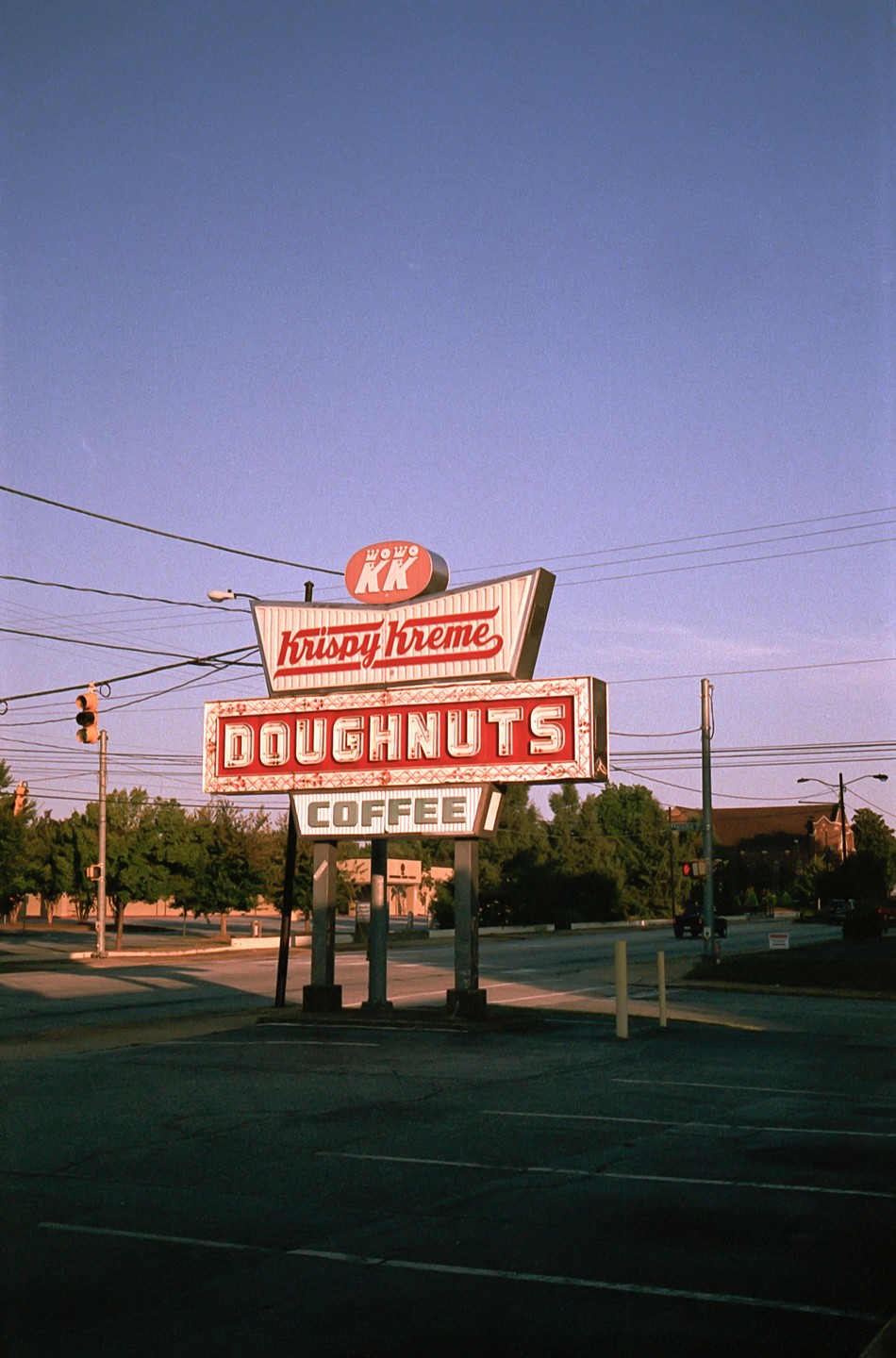 A Krispy Kreme sign stands at a street corner with trees and buildings in the background.