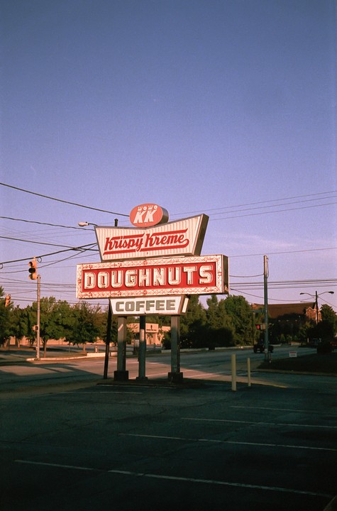A Krispy Kreme sign stands at a street corner with trees and buildings in the background.
