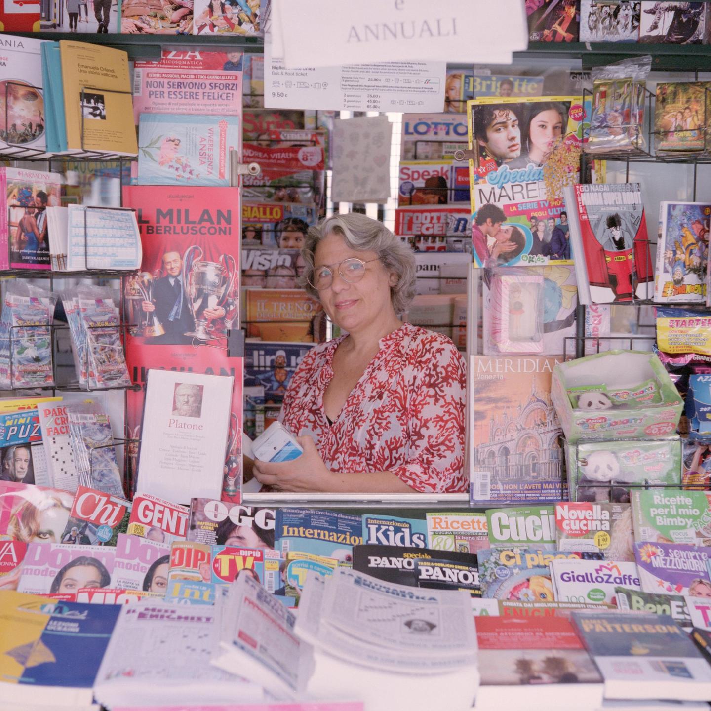 A woman with glasses and a patterned shirt stands behind a counter filled with magazines and newspapers.